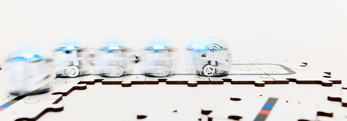 Kleiner, runder, leuchtender Roboter mit kleinen Rädern fährt auf Puzzleteilen, die Informationen in Form von schwarzen Linien und farbigen Codes bereithalten.