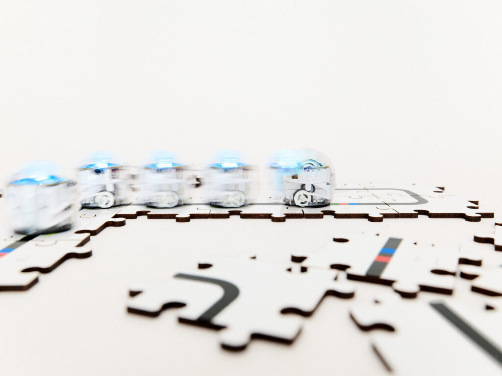 Kleine, runde Roboter mit kleinen Rädern fahren auf Matte, die Informationen in Form von schwarzen Linien und farbigen Codes bereithält.